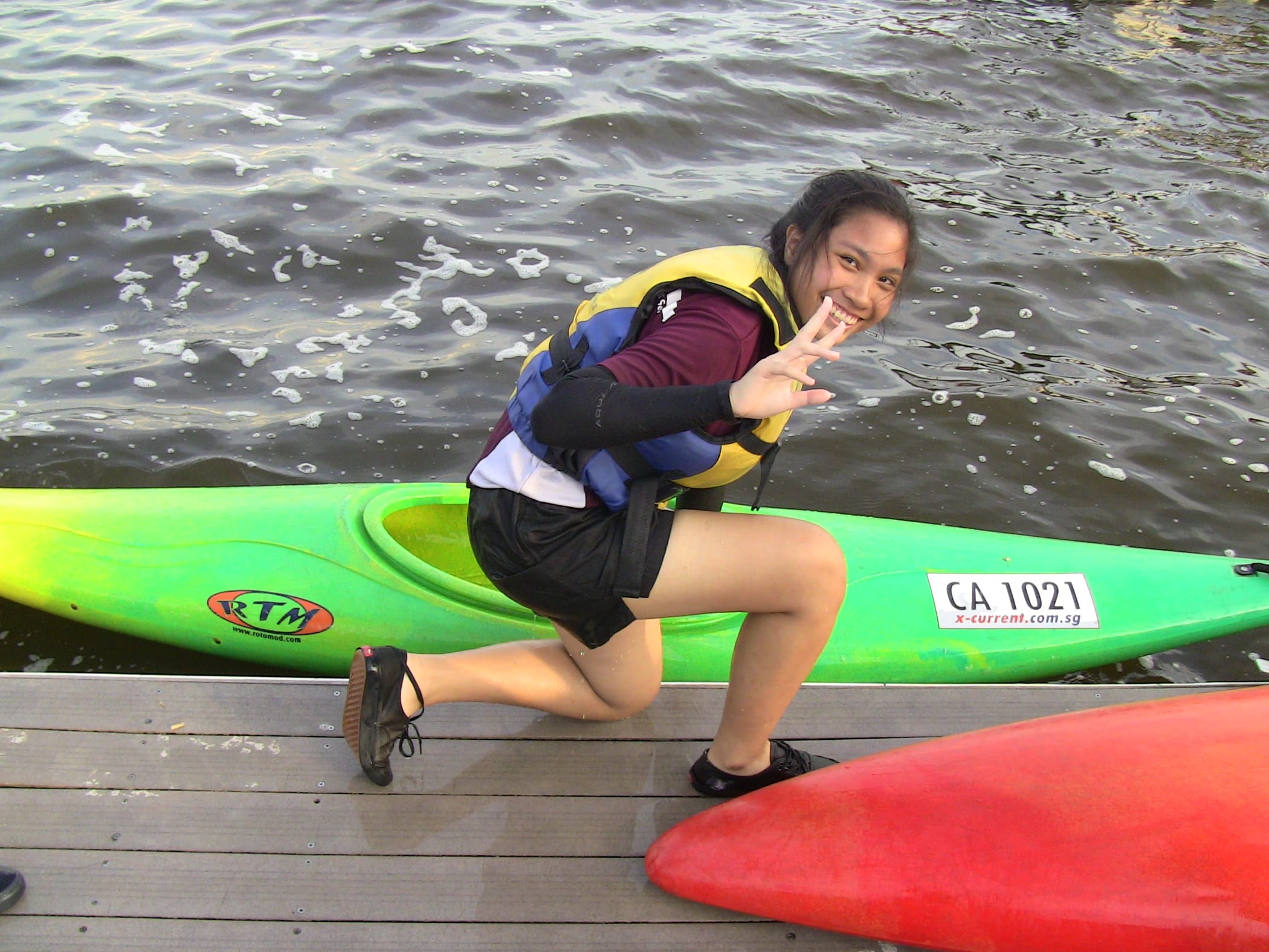 Young woman in a yellow life vest poses with a peace sign beside a green RTM kayak on a wooden dock.